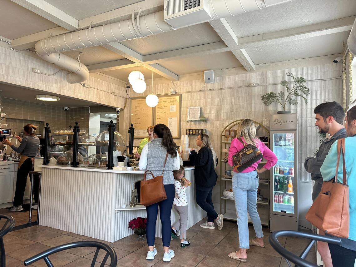 The interior of a busy coffee shop. Customers stand in line at a white, textured counter, waiting to order.