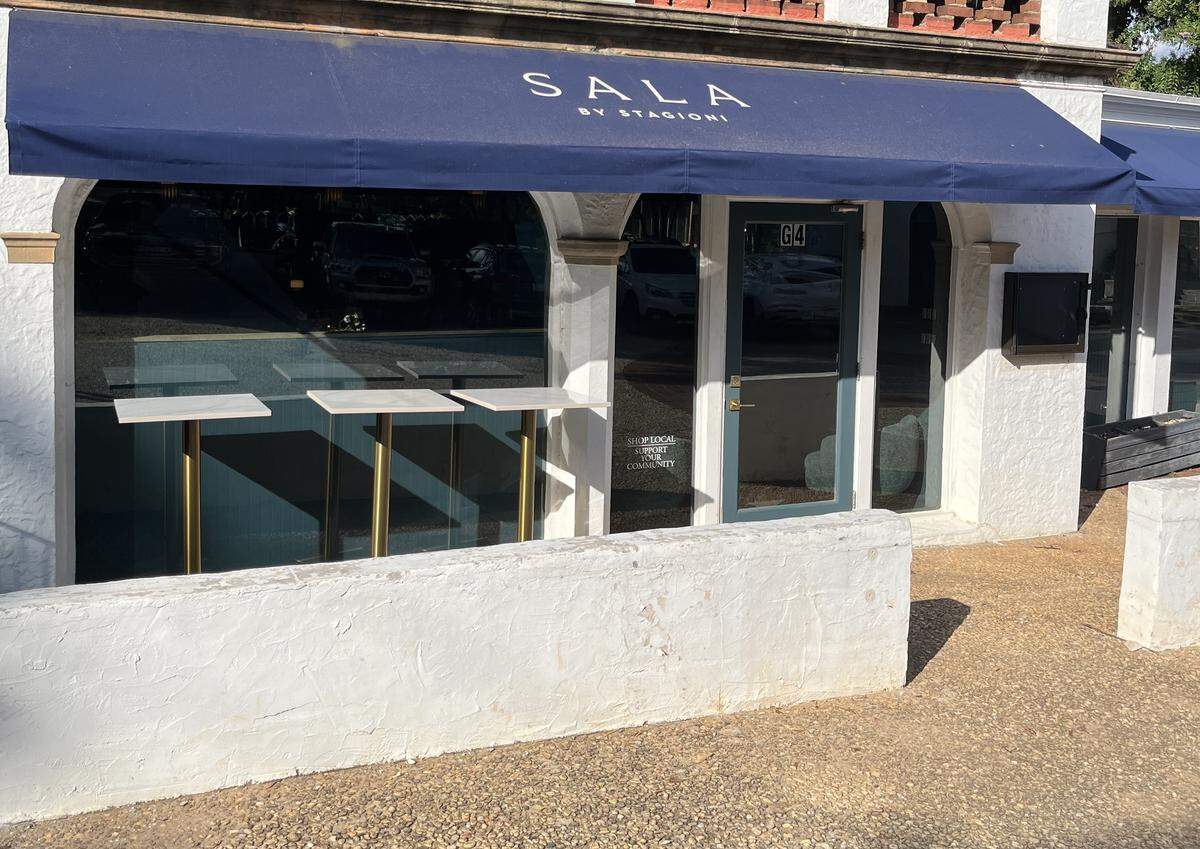 A wide shot of the entrance to Sala by Stagioni restaurant. A low, white stucco wall is in the foreground. The building itself has a matching stucco exterior with large, arched windows and a glass door. A deep blue awning extends over the storefront, with the restaurant’s name printed in white.