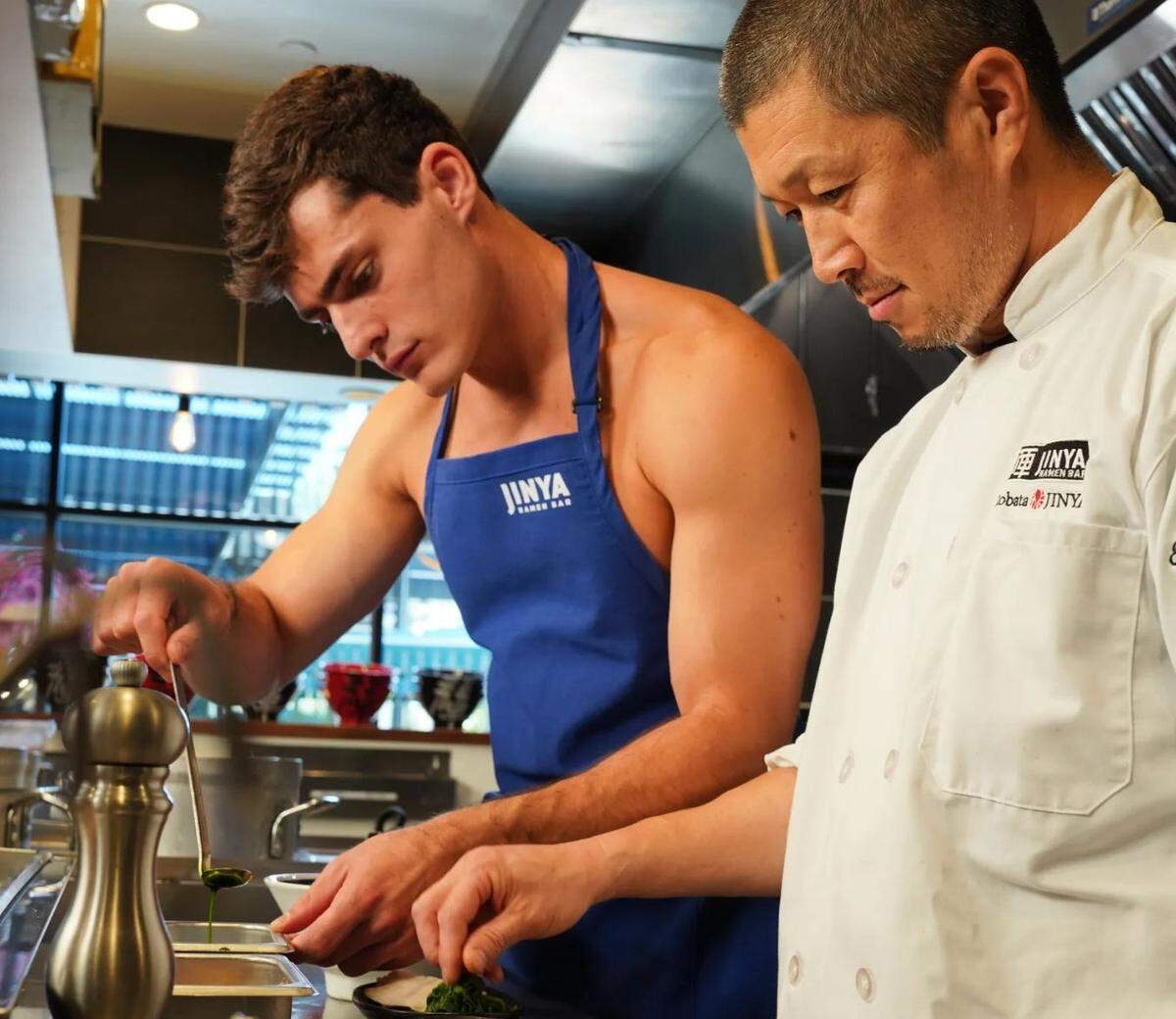 Two chefs are focused on preparing a dish in a professional kitchen. The chef on the left is a young man with a dark apron and no shirt, meticulously using a spoon to drizzle a green sauce onto a small bowl. The older chef on the right, wearing a white chef’s jacket with a “JINYA Ramen Bar” logo, looks on intently, assisting with the preparation. They are standing behind a stainless steel counter with various kitchen equipment.
