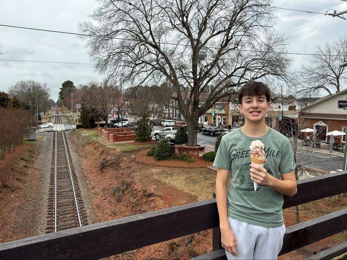 A young person standing on a wooden pedestrian bridge overlooking a long, straight set of railroad tracks while holding a waffle cone with sprinkles. The background shows the small-town charm of downtown Waxhaw under an overcast sky.