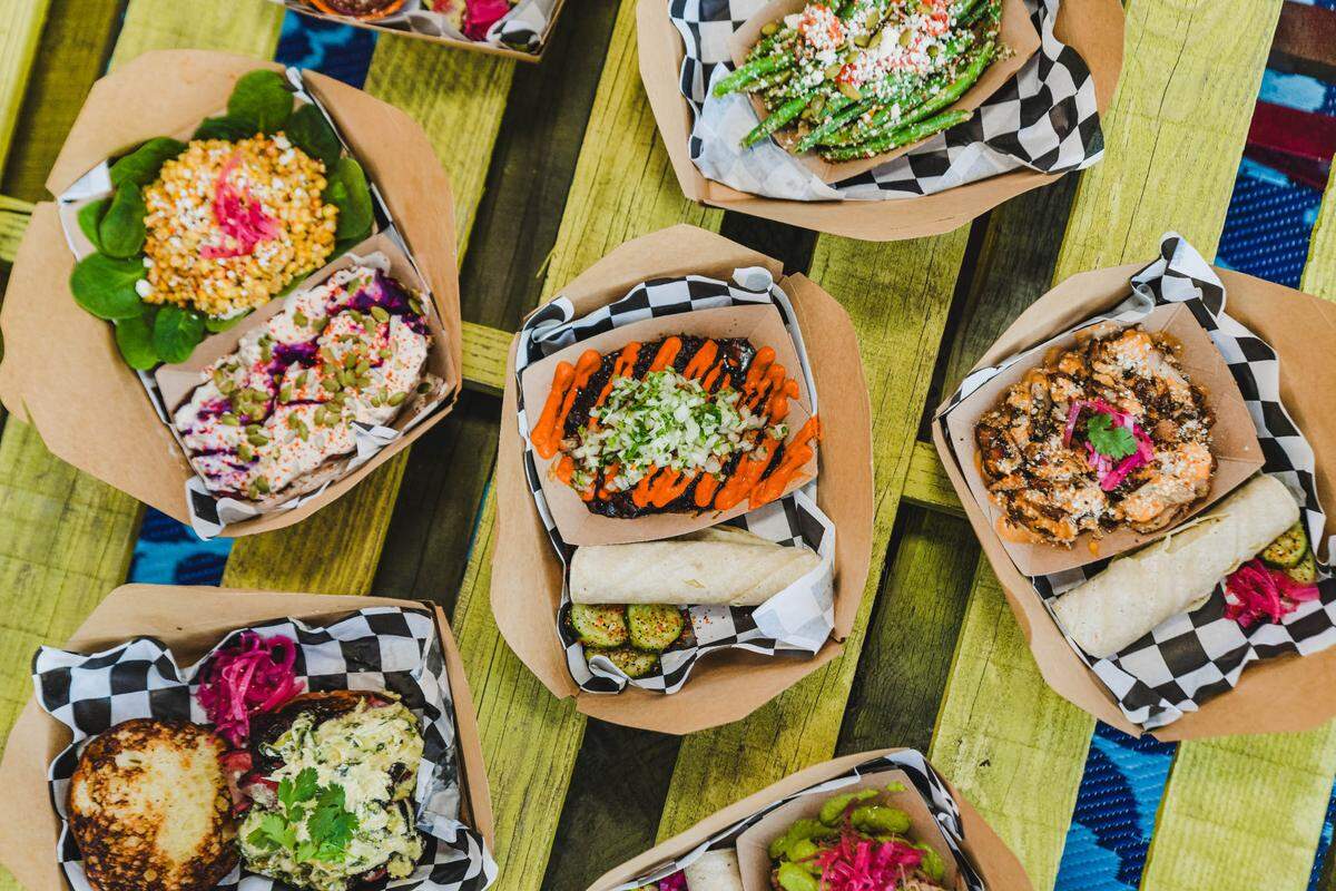 A vibrant top-down shot of a variety of street food dishes, likely from a food truck or market. Several cardboard takeout trays, each lined with black and white checkered paper, are arranged on a rustic light green wooden surface. Dishes include a black bean-based dish with orange and white sauce, a salad with pink and yellow grains, green beans topped with cheese and red flakes, and a dish with grilled chicken or fish accompanied by tortillas and pickled red onions. Other trays show sandwiches and various colorful toppings and garnishes.