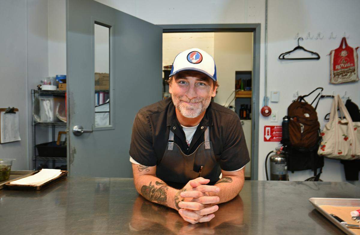 A chef with a beard, tattoos and a Grateful Dead baseball cap, smiles warmly while leaning on a stainless steel counter in what appears to be a commercial kitchen. The chef is wearing a dark shirt and an apron. In the background, there is a doorway, a fire extinguisher, and bags hanging on hooks.