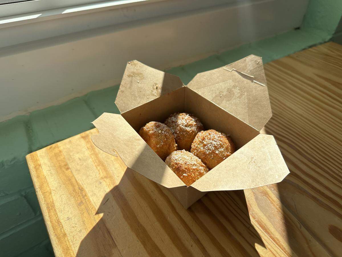 A top-down view of four golden-brown fried rice balls nestled inside a brown cardboard takeout box. The box sits on a light wooden surface in a bright, sunlit area, creating a strong diagonal shadow across the wood.