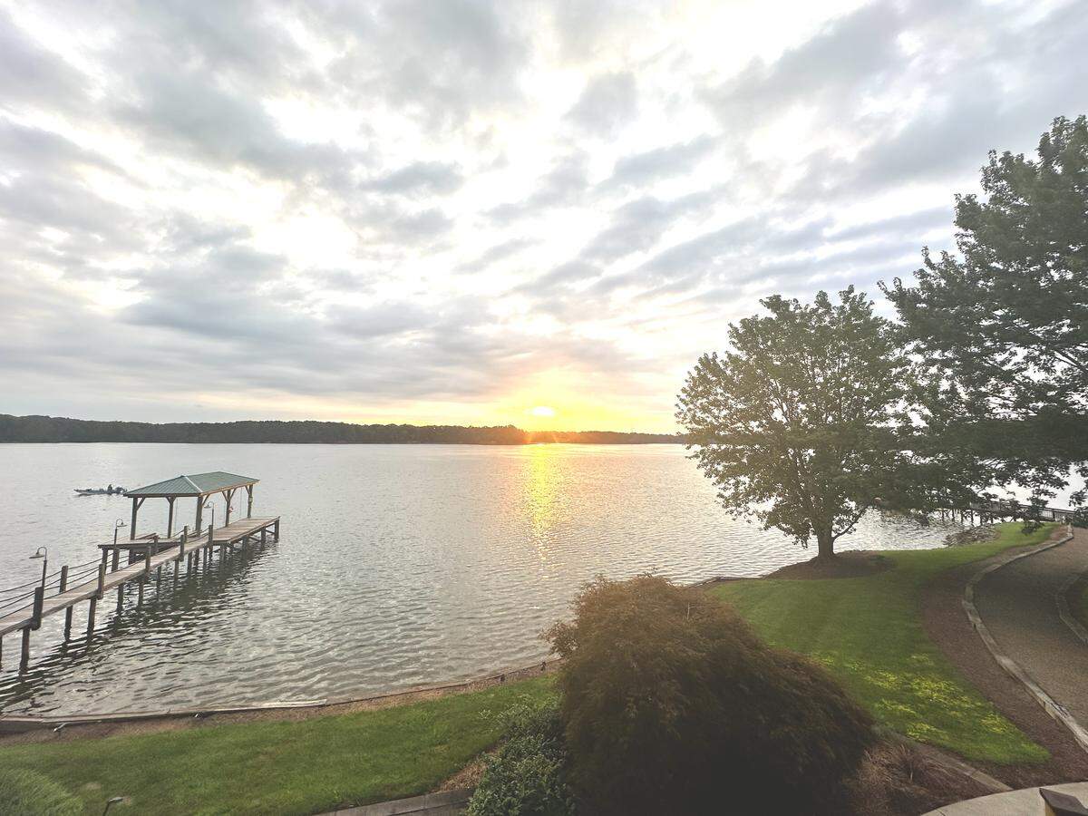 A scenic view of Lake Norman with the sun on the horizon, showing two wooden docks along a grassy shoreline. One dock with a green roof is prominent in the foreground, while a second dock is visible in the distance behind some trees. The sun creates a bright reflection across the water.