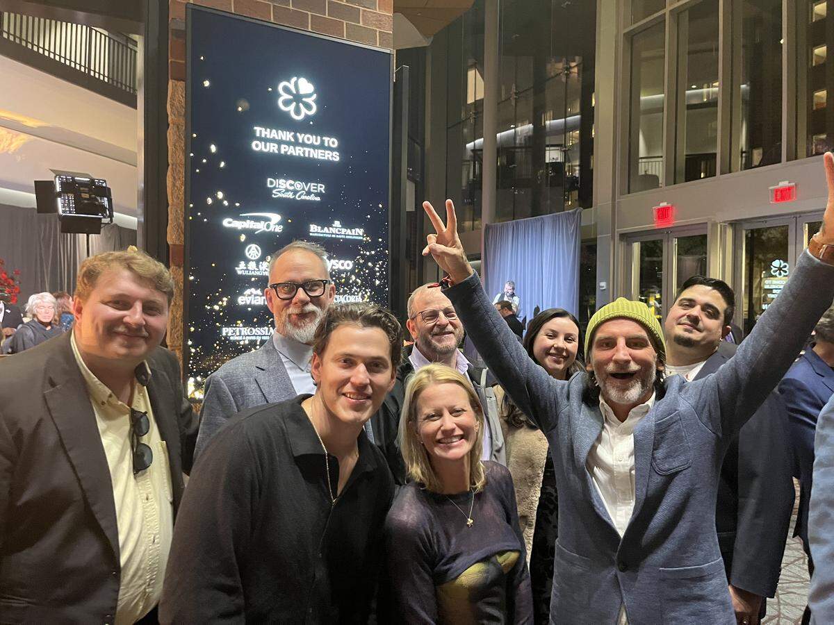 A group of eight people poses for a photo at an indoor evening event, in front of a tall black digital sign that reads “THANK YOU TO OUR PARTNERS.” The person in the front right, wearing a blue blazer and a lime green beanie, is smiling with arms raised, making a peace sign with one hand. A person with blonde hair stands next to that beanie-wearing person in the center, and several others, all smiling, are grouped closely around them.