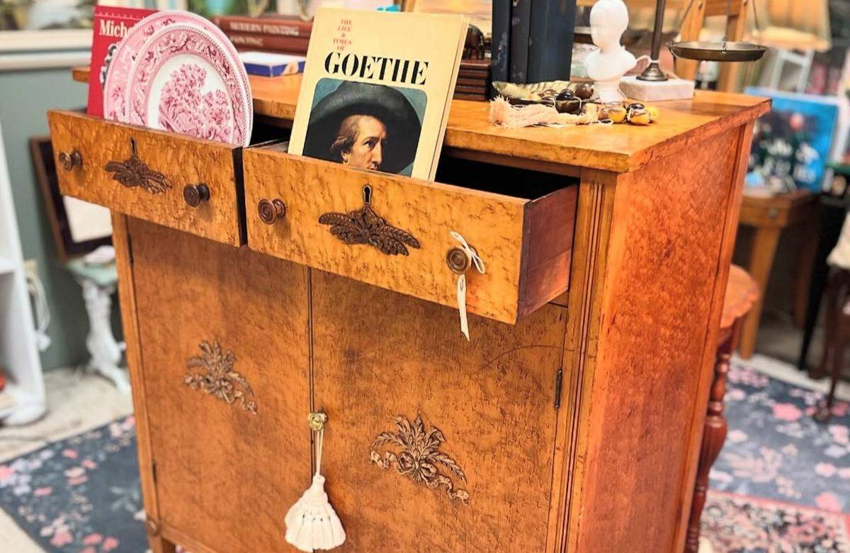 A close-up, angled view of an antique cabinet made from honey-colored bird’s-eye maple. The two upper drawers are open, showcasing items for sale. On top of the cabinet, a collection of antiques, including a set of scales and a small white bust, are arranged. The cabinet has decorative dark metal hardware and a key with a tag in the lock.