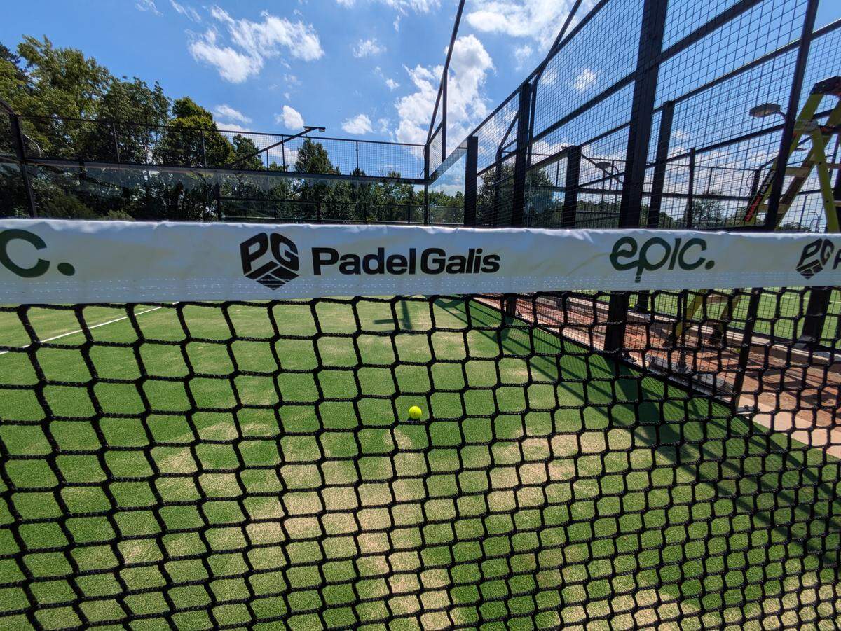 A low-angle shot from the net of a padel court on a sunny day. The white net tape is in the foreground, and through the mesh, a single yellow padel ball is visible on the green turf. In the background are the court’s fence, trees, and a blue sky with clouds. A ladder is visible on the right.