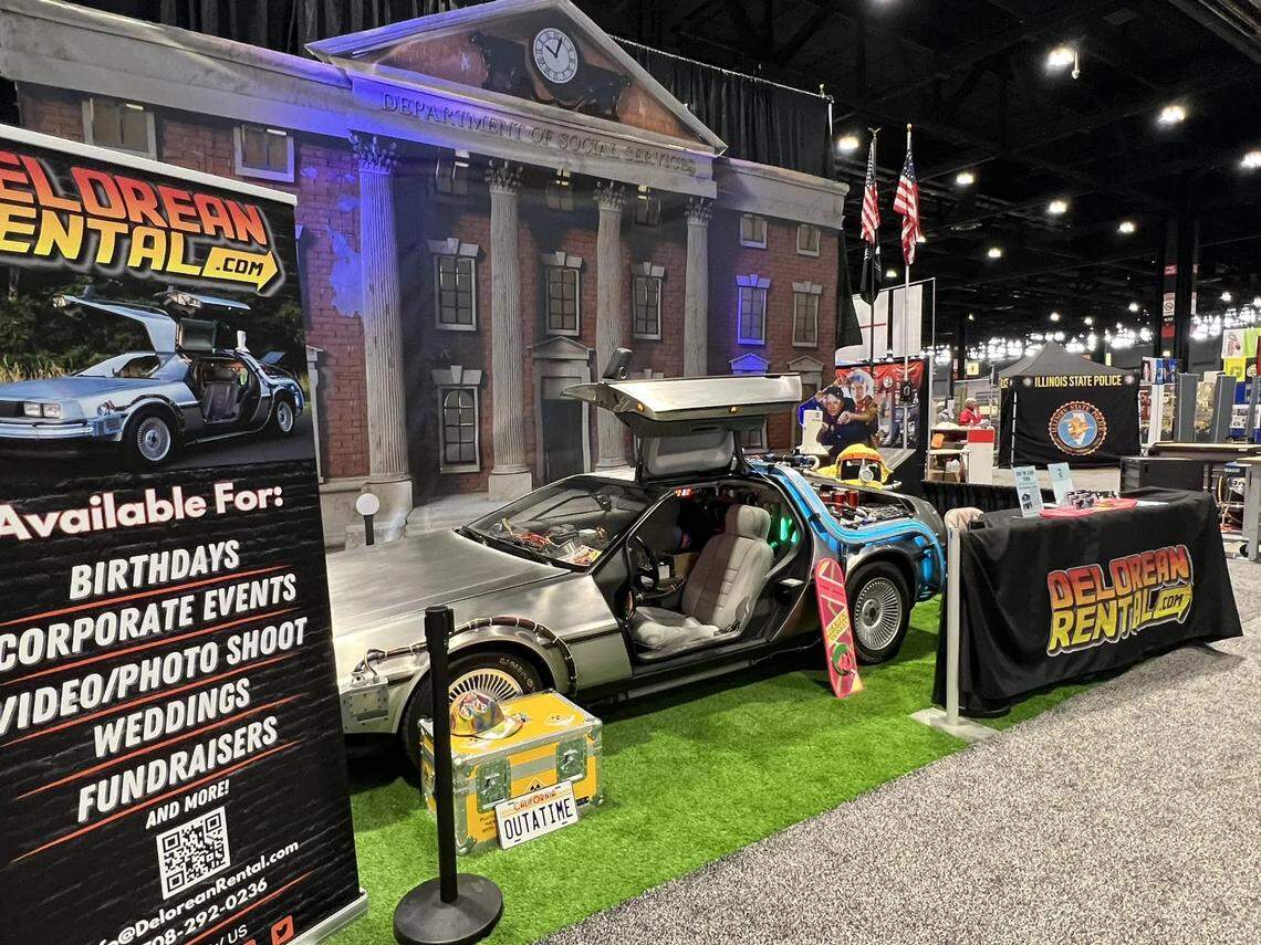 A wide shot of a “Back to the Future” exhibit at a convention or trade show, featuring a DeLorean time machine replica. The car has its gull-wing doors open and is parked on a patch of artificial turf in front of a large backdrop depicting the Hill Valley clock tower. A pink hoverboard leans against the car. To the left, a large banner advertises “DELOREAN RENTAL.COM” for various events. A yellow prop “OUTATIME” license plate case sits in front of the car, and a table to the right, draped in black, also features the rental company’s logo.