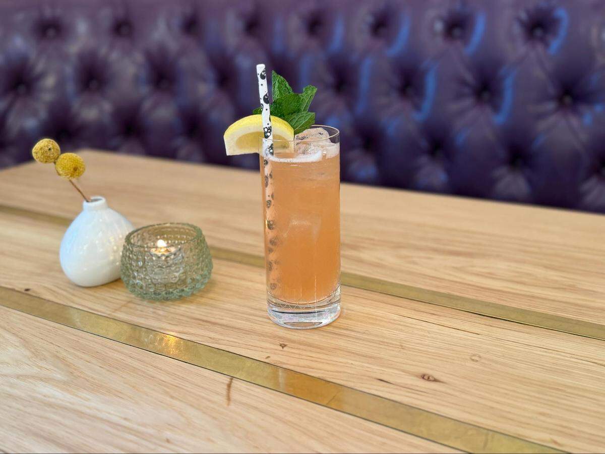 A close-up, eye-level shot of a tall, light-orange cocktail on a light wooden table with brass inlays. The drink is in a highball glass and is garnished with a fresh mint sprig, a lemon wedge, and a decorative paper straw. In the background, a purple, tufted booth is visible, and to the left of the glass sit a small candle holder and a white vase with yellow flowers.