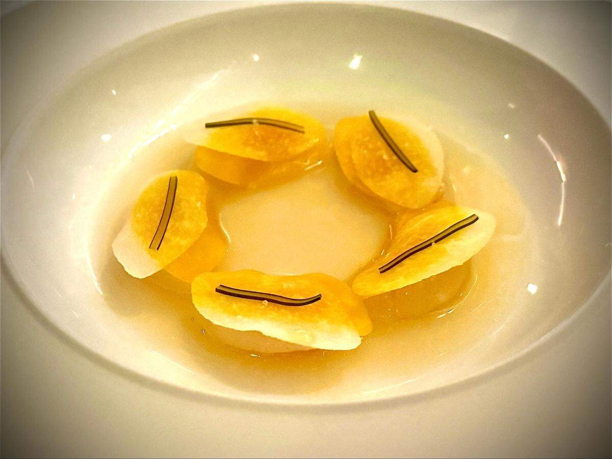 A close-up, high-angle shot of a white, shallow bowl containing a clear, amber-colored broth. Floating in the broth are about six artfully arranged, petal-shaped slices of what appear to be poached scallops, each garnished with grapefruit and a tiny, dark green sprig of herbs.