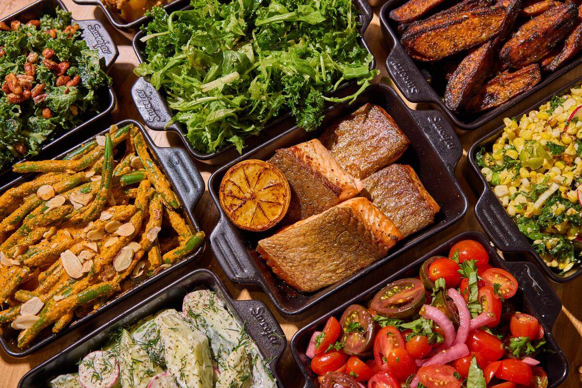 A close-up, angled view of several rectangular black serving dishes filled with assorted colorful foods on a wooden surface. In the center, four pieces of golden-brown seared salmon are served with a grilled lemon half. Surrounding the salmon are dishes containing a bright tomato and red onion salad, creamy cucumber salad with dill, roasted potato wedges, sautéed green beans with almond slivers, and a corn and kale salad. The lighting is warm and bright, emphasizing the textures of the fresh ingredients.