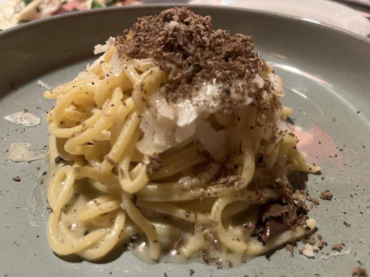 A close-up, high-angle shot of a gourmet pasta dish served in a matte grey bowl. The dish features a swirl of thick, long pasta—likely tonnarelli—coated in a creamy, light-colored sauce. It is generously topped with a mountain of finely shaved black truffles and flakes of Pecorino or Parmesan cheese. Speckles of cracked black pepper are scattered across the pasta and the rim of the bowl, highlighting the rich, earthy textures of the meal.