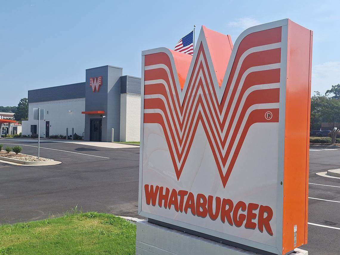 A large, white and orange striped “W” logo stands prominently in the foreground on a brick pedestal in front of a Whataburger restaurant. The modern building features a gray and white exterior with an American flag flying in the background under a clear blue sky.