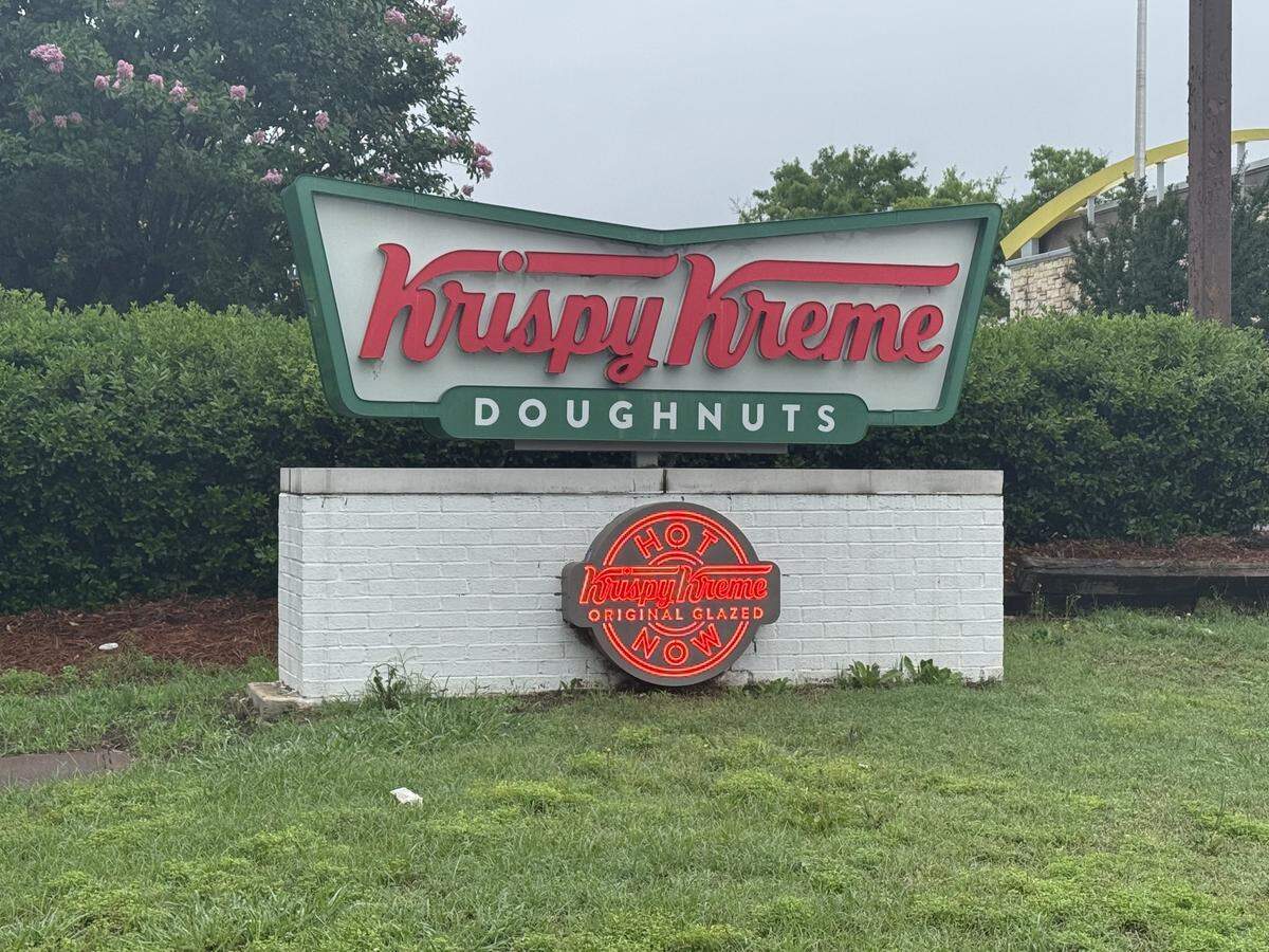 A street-level roadside sign for Krispy Kreme Doughnuts on a cloudy day. The top portion features the classic retro green bowtie-shaped sign with red cursive text reading “Krispy Kreme” above a green bar that reads “DOUGHNUTS” in white capital letters. The sign is mounted on a low, white brick base. Set into the center of the brick base is the circular “Hot Light” neon sign, which is illuminated in bright red and reads “HOT Krispy Kreme ORIGINAL GLAZED NOW”. Green grass sits in the foreground, and a dense green hedge with a few pink flowers lines the background.