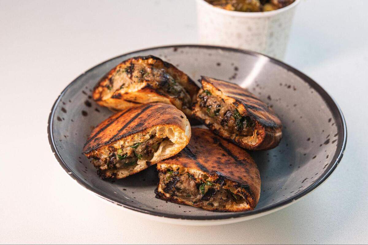 A high-angle shot of a gray bowl with four pieces of grilled, triangle-shaped flatbread filled with a savory mixture of meat and herbs. The flatbread has prominent char marks from grilling. In the blurred background, a small container of olives is visible. The bowl rests on a white surface.