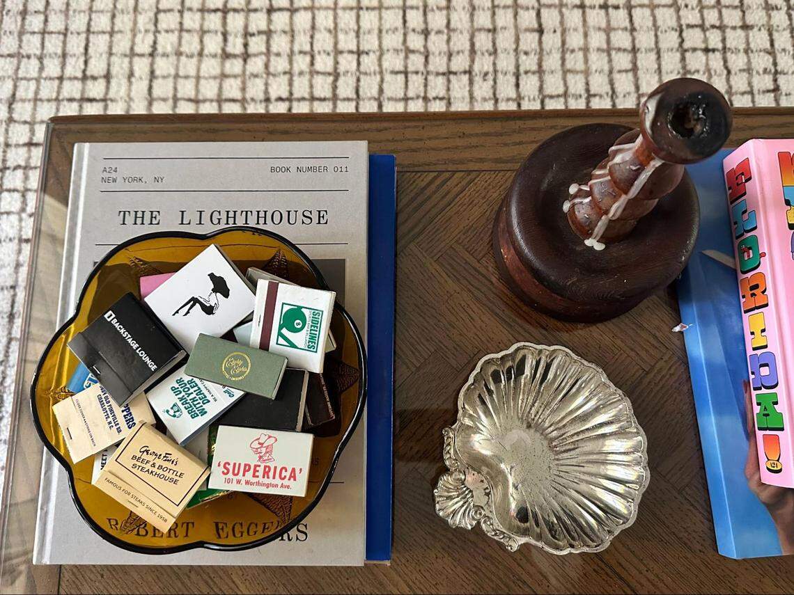 An overhead shot of a styled wooden coffee table. The central focus is a yellow, flower-shaped glass bowl filled with a collection of matchbooks from various establishments, including “BACKSTAGE LOUNGE,” “SUPERICA,” and “Gene & Georgetti STEAKHOUSE.” The bowl sits atop a book titled “THE LIGHTHOUSE.” To the right of the bowl is a small, silver, shell-shaped dish and a dark wooden object that looks like a wax seal stamp. A stack of colorful books is visible on the far right, and the table is placed on a light-colored, patterned rug.