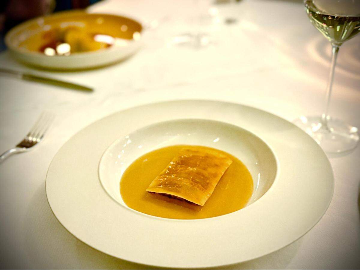 A high-angle, close-up shot of a single, large, rectangular ravioli presented in the center of a wide, white-rimmed bowl. The ravioli is floating in a smooth, amber-colored broth that fills the bottom of the bowl. The dish sits on a white tablecloth, with a fork and a wine glass partially visible in the blurred background.