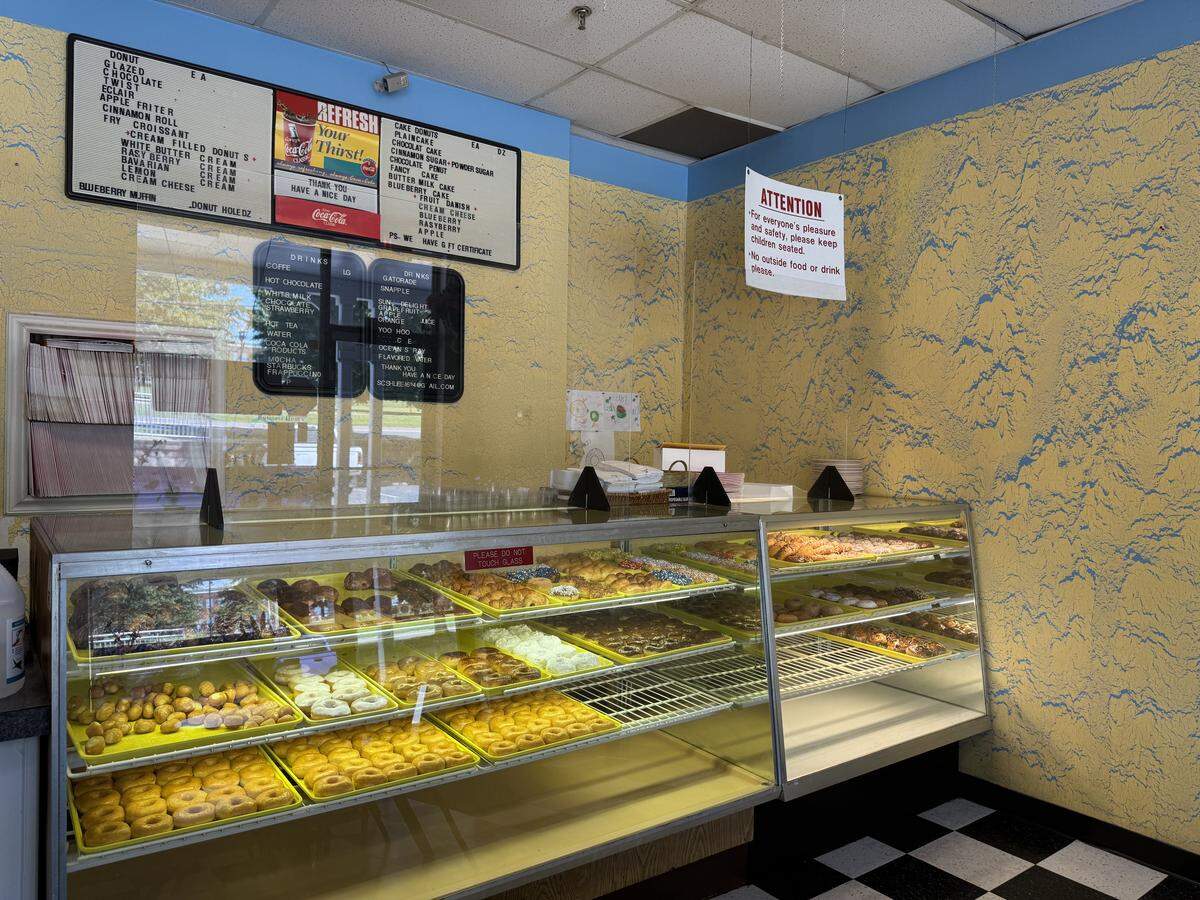 A complete view of the service area of an old-fashioned donut shop. A multi-level glass display case is filled with a wide assortment of donuts. The decor includes a black and white tile floor and yellow walls with a blue pattern. Above the counter, several menu boards list the available donuts and drinks.