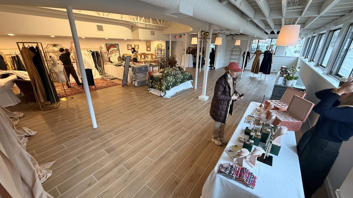 A wide-angle interior shot of a brightly lit, modern event space with light wood plank flooring. Vendor tables, draped in white and featuring jewelry and clothing displays, line the right side of the room. A woman in a brown jacket and a red cap is looking at a table. In the background, clothing racks and mannequins stand near large windows, and an attendant is working near a clothing display on the left.