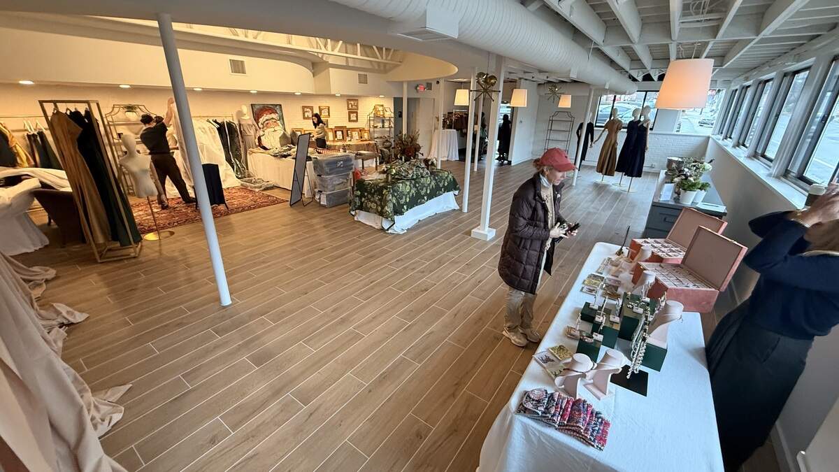 A wide-angle interior shot of a brightly lit, modern event space with light wood plank flooring. Vendor tables, draped in white and featuring jewelry and clothing displays, line the right side of the room. A woman in a brown jacket and a red cap is looking at a table. In the background, clothing racks and mannequins stand near large windows, and an attendant is working near a clothing display on the left.
