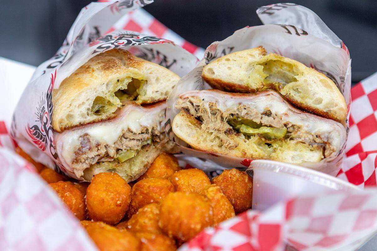 A professional close-up of a meal served in a basket lined with red and white checkered paper. A crusty roll sandwich is sliced in two, displaying its generous filling of shredded meat, melted provolone-style cheese, and pickles. A pile of crispy, round tater tots fills the front of the basket.