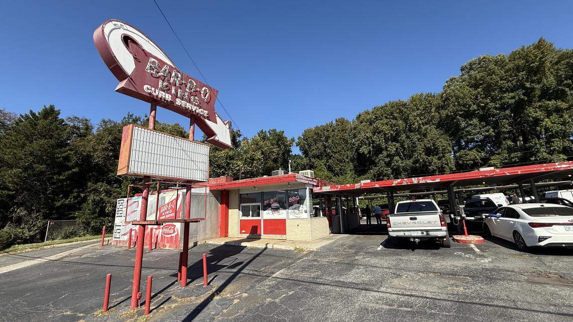 A wide, eye-level shot of the “Bar-B-Q King,” a vintage drive-in restaurant, on a sunny day. A large, retro red and white sign with a curved arrow advertises “BAR-B-Q KING” and “CURB SERVICE.” The small red and white building has a large red canopy extending over the parking lot, where several cars are parked. The scene is set against a backdrop of dense green trees and a clear blue sky.