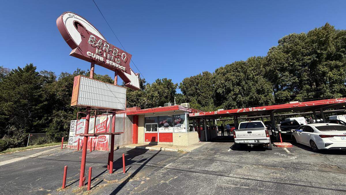 A wide, eye-level shot of the “Bar-B-Q King,” a vintage drive-in restaurant, on a sunny day. A large, retro red and white sign with a curved arrow advertises “BAR-B-Q KING” and “CURB SERVICE.” The small red and white building has a large red canopy extending over the parking lot, where several cars are parked. The scene is set against a backdrop of dense green trees and a clear blue sky.
