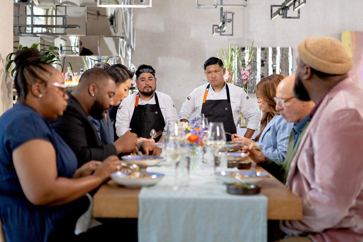 A group of people are seated at a long, wooden dining table in a bright, modern restaurant, tasting various dishes. Two chefs in white coats and black aprons stand at the head of the table, attentively watching the diners’ reactions. The table is set with a light blue runner, wine glasses, and floral arrangements, while the background features a unique wall of geometric mirrors.