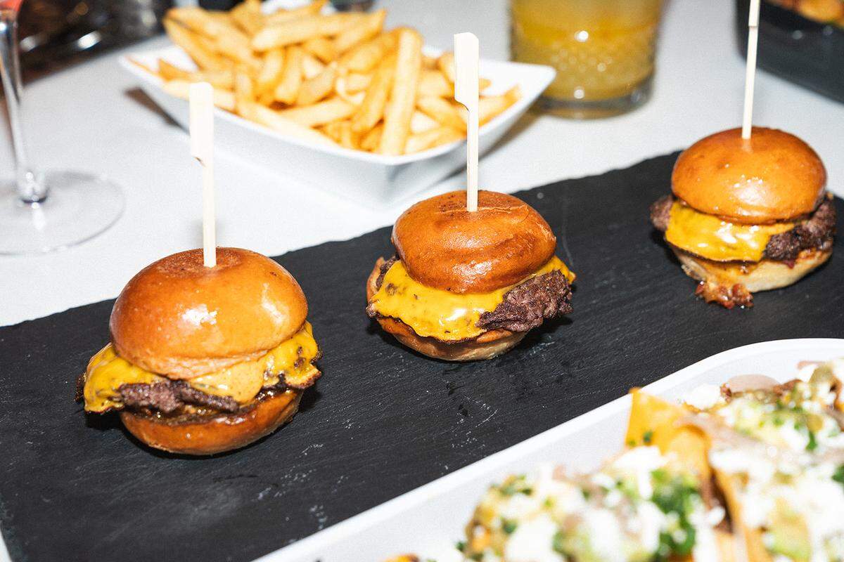 A food spread at the Blinders bar is shown in a close-up shot. The focus is on three cheeseburger sliders on a slate platter, each held together with a white pick. A white bowl of fries is in the background, and another dish with toppings is in the foreground, creating a scene of a shared meal.