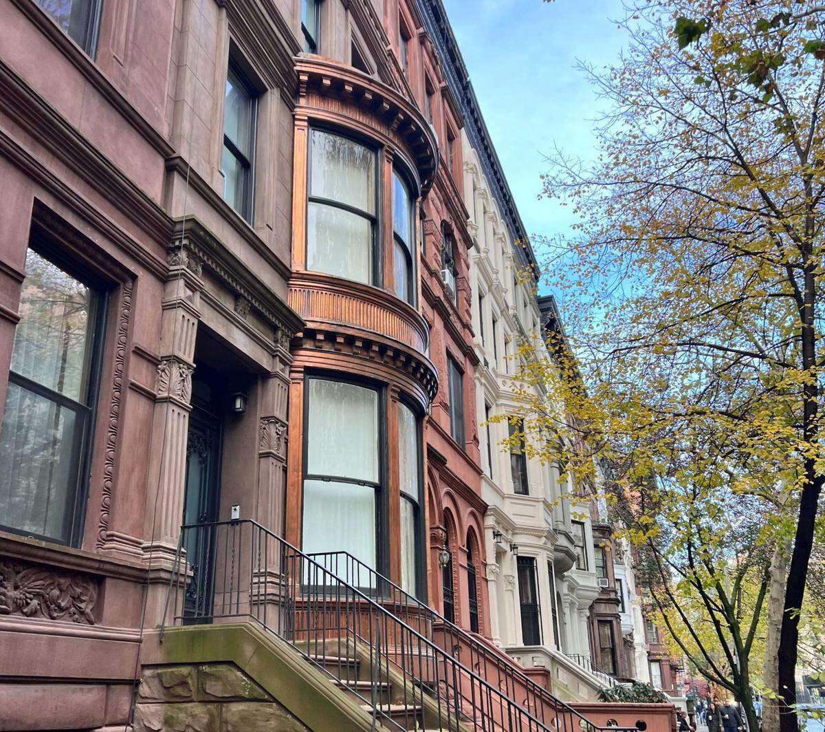 A close-up, angled shot of a row of classic brownstone buildings in a city neighborhood. The buildings are made of reddish-brown stone, each with a stoop and a staircase leading to the entrance. The building in the center has a prominent rounded bay window. A tree with yellow autumn leaves is in the foreground on the right, and the sky is a bright blue with some clouds.