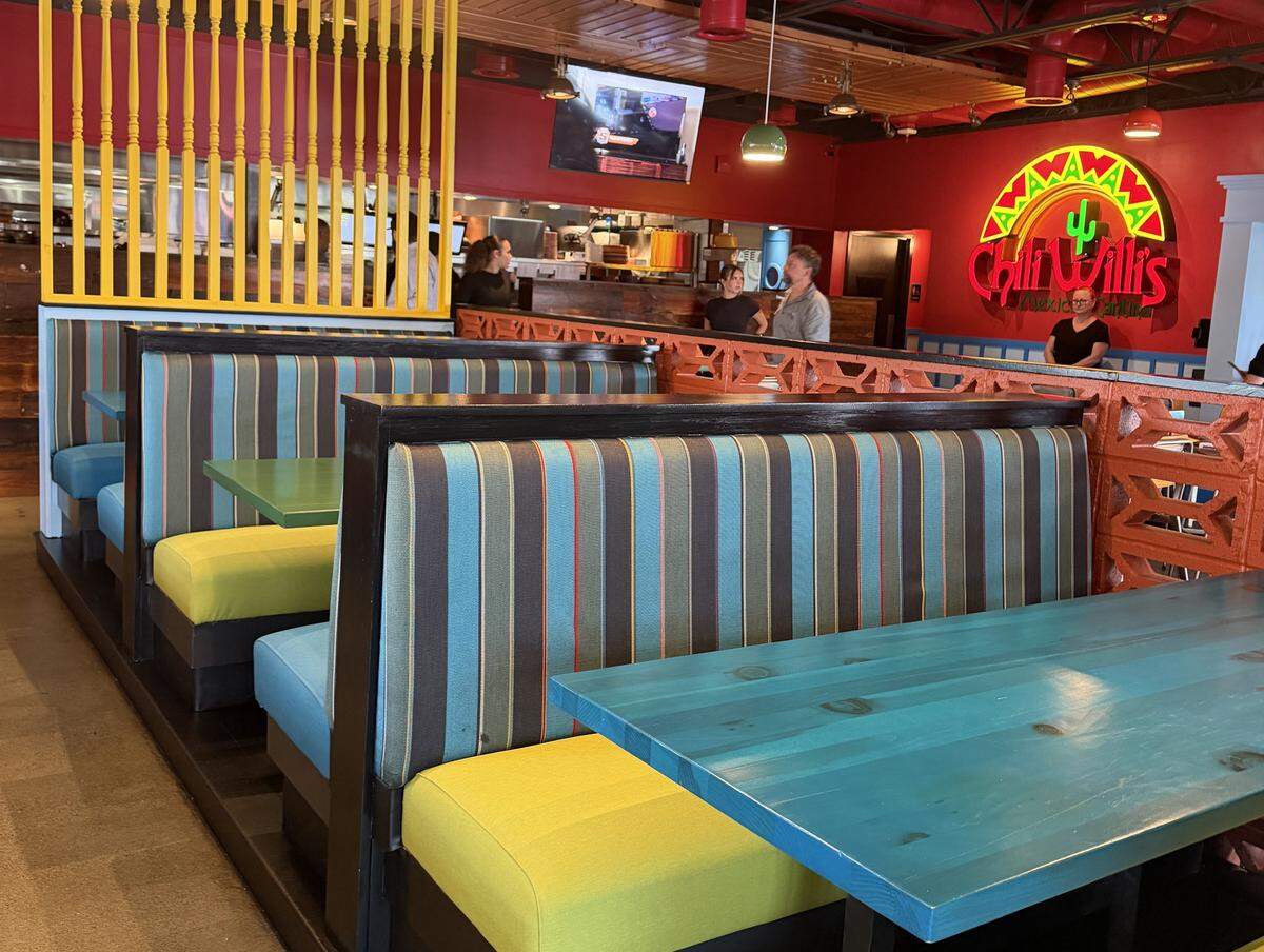 A row of restaurant booths with striped upholstery in teal, brown, and red, paired with bright lime-yellow seat cushions. The tables are a vibrant teal wood. A decorative orange cinderblock wall and a bright yellow spindled divider separate the seating areas.