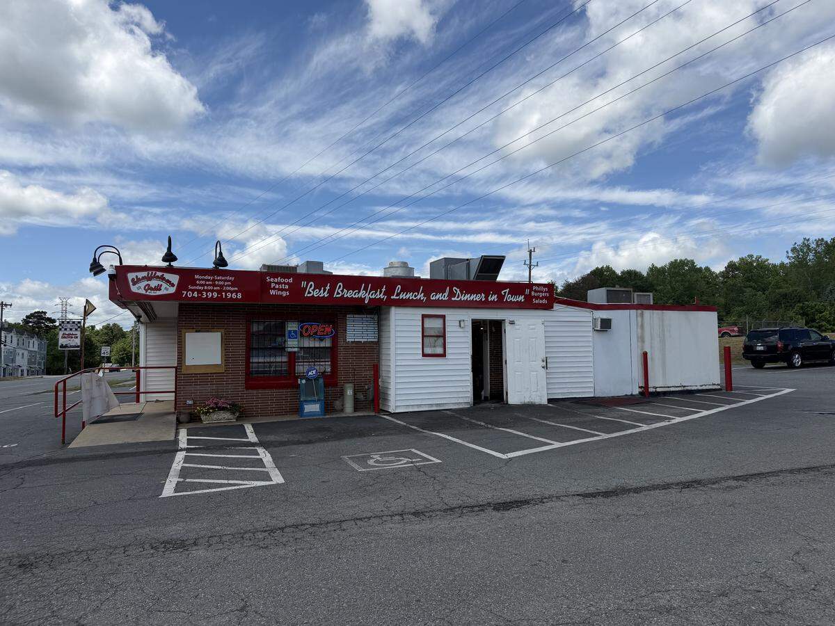 A side view of Shuffletown Grill featuring a red awning with the restaurant’s logo, phone number, and operating hours. The building is a mix of red brick and white wood siding. A red metal railing leads to the entrance, and an “Open” neon sign is visible in the window. The parking lot has several marked spaces, including a designated handicap spot.