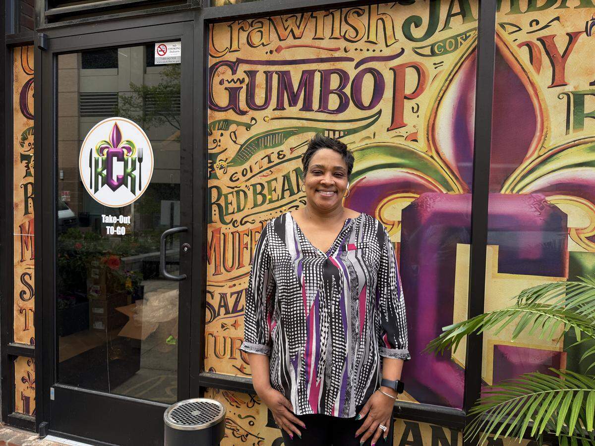 A smiling person with short dark hair and a warm, welcoming expression stands proudly in front of the window of Kristy’s Creole Kitchen (KCK), which is adorned with vibrant, colorful vinyl graphics.