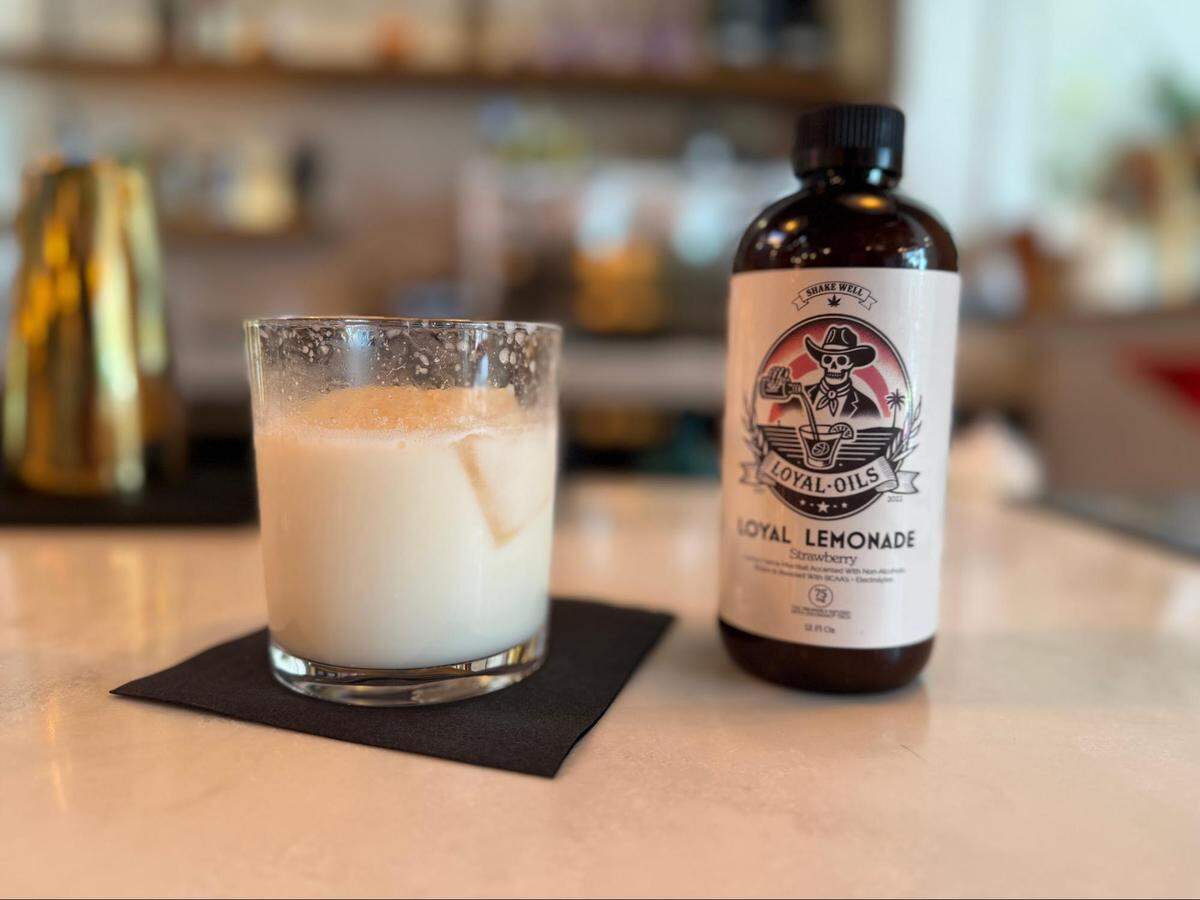 A close-up, eye-level shot of a creamy white cocktail in a salt-rimmed rocks glass, sitting on a black napkin. To its right is a bottle of “Loyal Oils Loyal Lemonade” strawberry mixer. The items are on a white and gray marbled bar top, with the rest of of the bar blurred in the background.