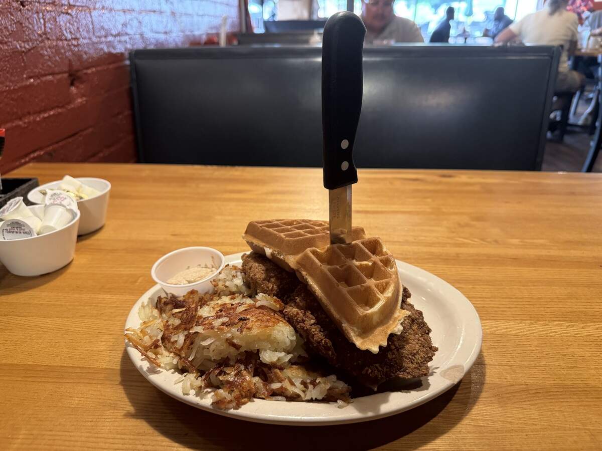 A close-up shot of a plate of chicken and waffles served on a light wooden table. A large, crispy fried chicken breast is topped with a golden-brown waffle section, held together by a black-handled steak knife stabbed vertically through the center. A generous side of crispy, browned hash browns and a small cup of butter sit on the white plate.