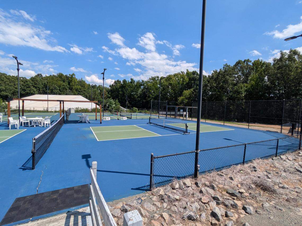 A wide-angle shot of an empty pickleball complex on a bright day. Several courts, painted blue with green non-volley zones, are shown. A covered seating area is visible on the left, and the entire facility is bordered by trees.