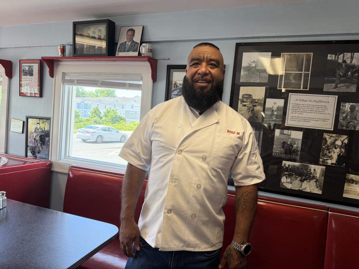 A portrait of a restaurateur with a full dark beard, identified by the red embroidery on his white double-breasted chef’s coat as “Jose M.” The person is smiling and standing in the dining area of Shuffletown Grill in front of a red vinyl booth and a wall display titled “A Tribute To Shuffletown,” featuring various historical black-and-white photographs and text about the local community. Through a window to the left, a bright day and several parked cars are visible.