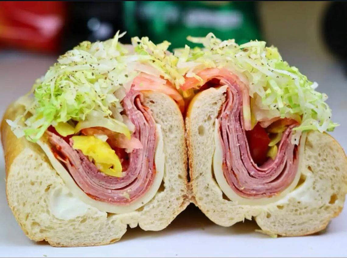 A close-up cross-section of a large sub sandwich cut in half. The bread is stuffed with a thick, U-shaped layer of folded Italian meats and white cheese. The center is packed with shredded iceberg lettuce, sliced tomatoes, and yellow peppers, all seasoned with black pepper and dried herbs.