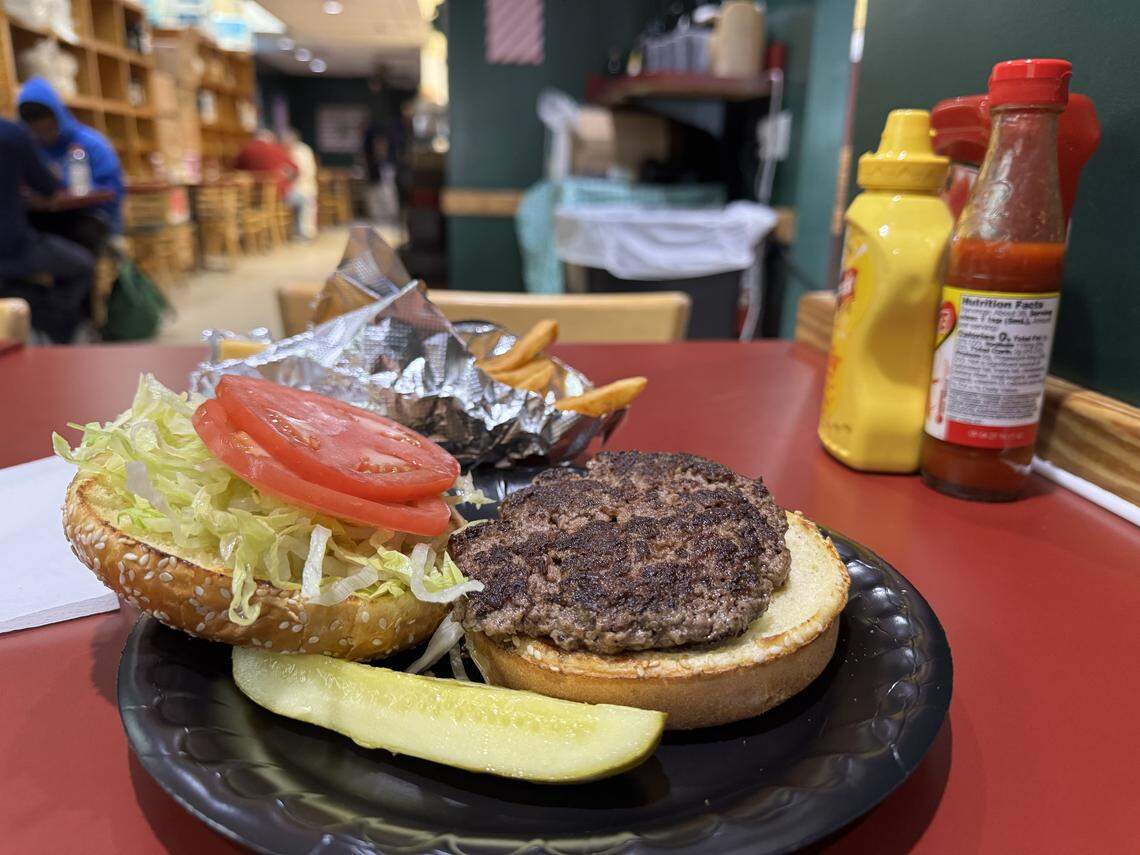 A close-up, eye-level shot of an open-faced hamburger served on a black disposable plate. The burger consists of a charred beef patty on a toasted sesame seed bun, with shredded lettuce and two thick tomato slices on the top half. A large pickle spear rests on the side. In the background, a small portion of French fries is visible in crumpled aluminum foil. Yellow mustard and red hot sauce bottles stand on the red tabletop.