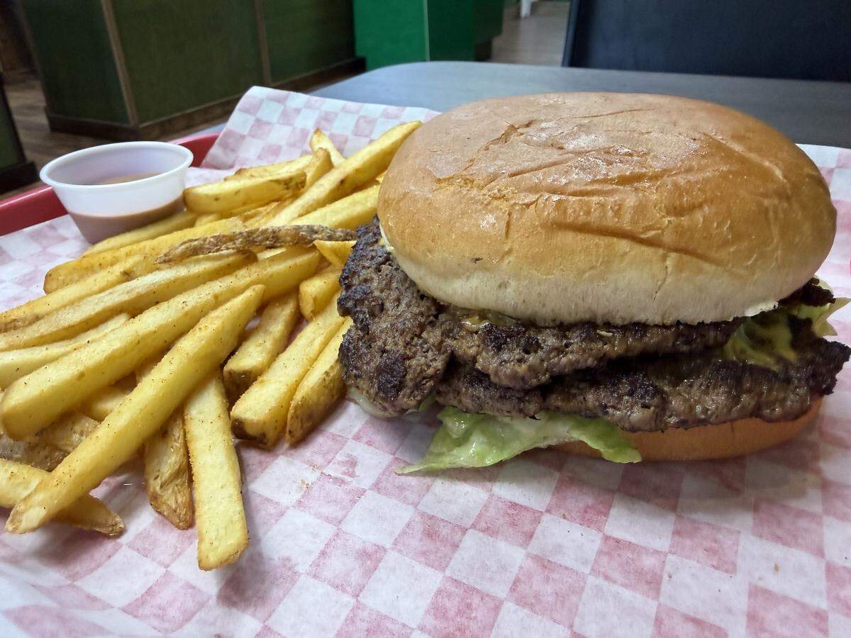 A close-up shot of a double cheeseburger and a side of french fries served on a red tray lined with pink and white checkered paper. The burger features two large, grilled beef patties stacked with melted cheese, lettuce, and sauce on a classic bun. A small plastic cup of dipping sauce sits next to the pile of golden, skin-on french fries.