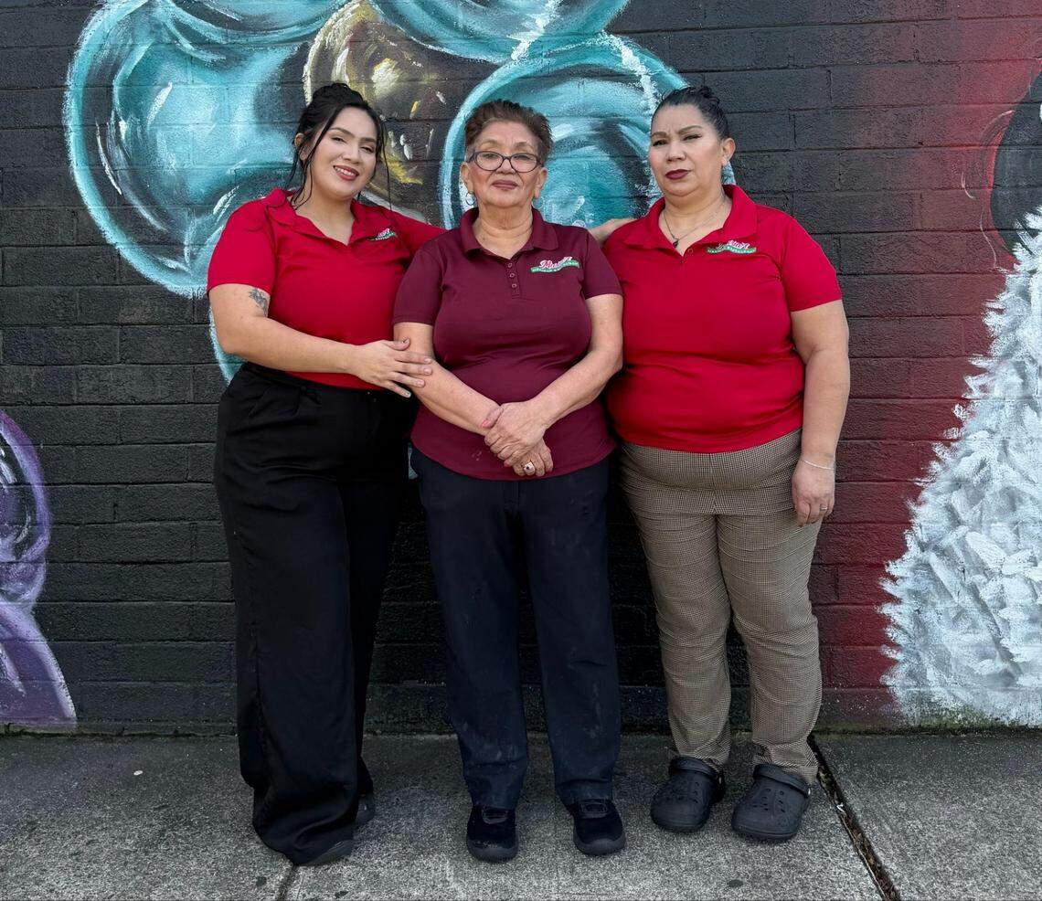 Three individuals stand together wearing matching red and maroon polo shirts in front of a dark brick wall featuring a large, colorful mural. Each person wears different styles of pants, including black, dark, and patterned options, while posing side-by-side.