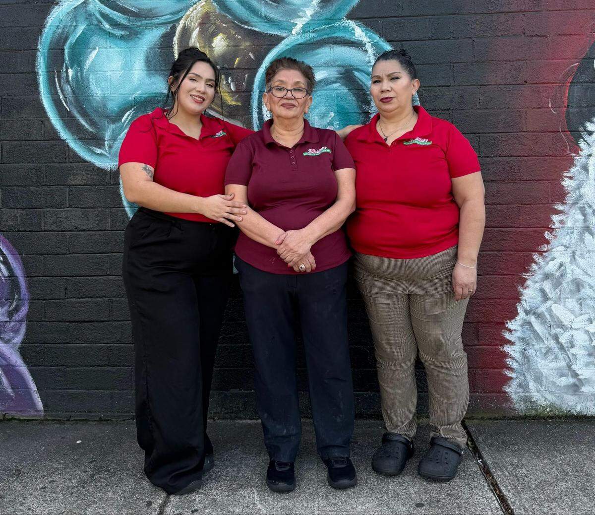 Three individuals stand together wearing matching red and maroon polo shirts in front of a dark brick wall featuring a large, colorful mural. Each person wears different styles of pants, including black, dark, and patterned options, while posing side-by-side.