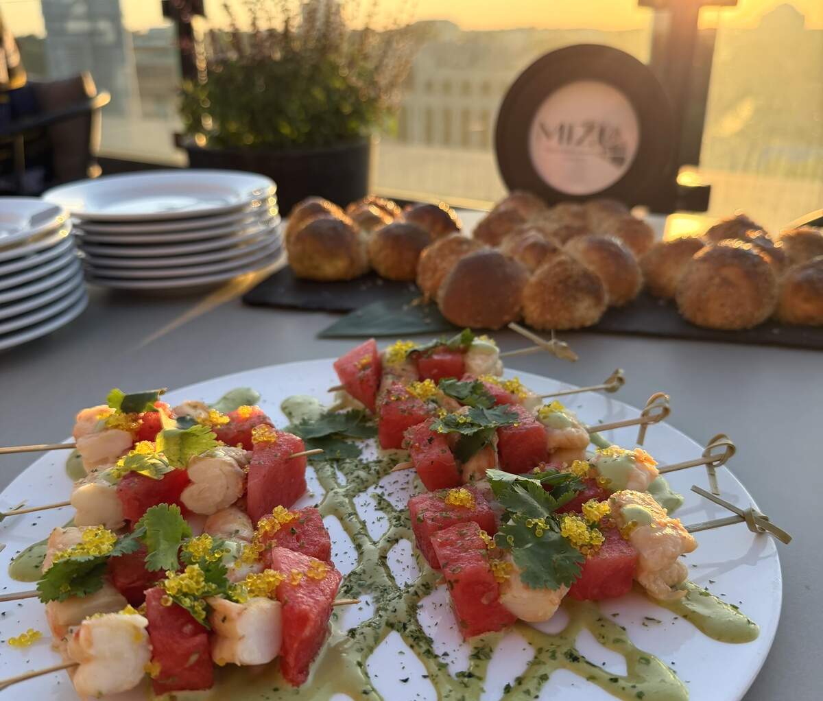 Skewers of cubed watermelon and shrimp garnished with yellow caviar and cilantro on a drizzled white plate, served on a rooftop table at Mizu during sunset with baked rolls in the background.