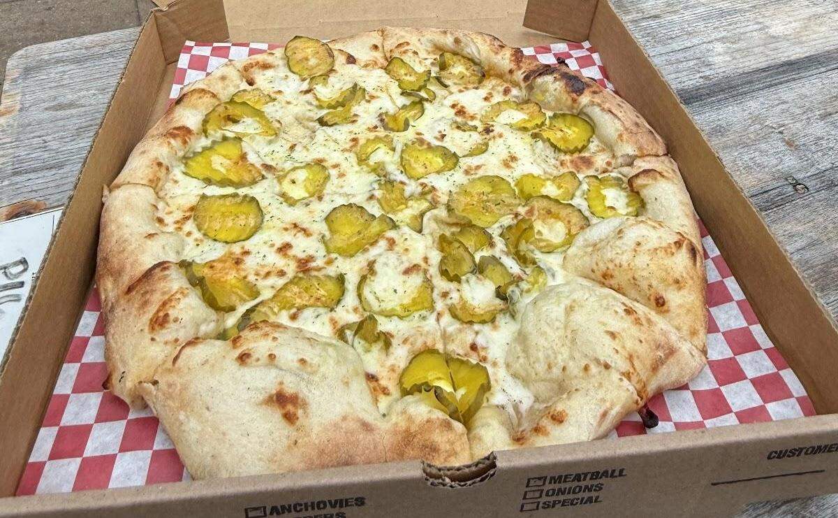 An overhead shot shows a whole pizza with a thick, hand-formed crust inside a takeout box with checkered paper. The pizza has a white sauce base and is topped with melted cheese and an abundance of sliced dill pickles, with a sprinkle of herbs.