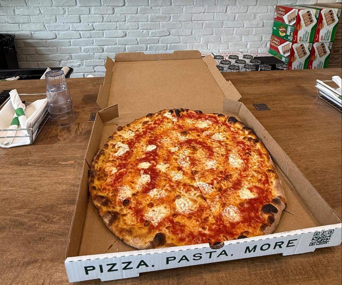 A high-angle, eye-level shot shows a large, round cheese pizza in an open “PIZZA, PASTA, MORE” cardboard box. The pizza, featuring red sauce and dollops of melted mozzarella, sits on a wooden counter, with a white brick wall and other counter items visible in the background.