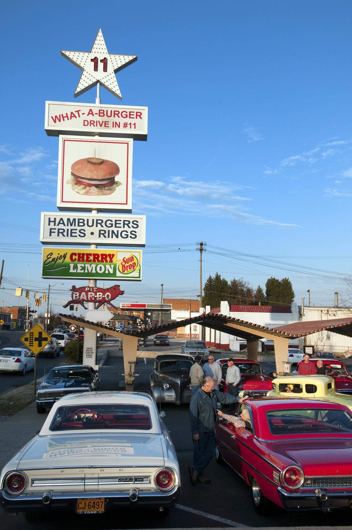 A daytime photo shows a bustling retro drive-in restaurant named “What-A-Burger Drive In #11” under a bright blue sky. A tall sign features a large white star with “11” on top, followed by the restaurant name, a picture of a hamburger, and text advertising “HAMBURGERS FRIES RINGS.” Below this, a green sign reads “Enjoy CHERRY Sun LEMON Drop.” In the foreground, several classic cars are parked, including a silver car facing away from the camera and a red car parked next to it, facing the right. Several people are gathered near the red car. In the background, more cars are visible, along with the restaurant building featuring covered drive-in stalls and additional signage for “BAR-B-Q.”