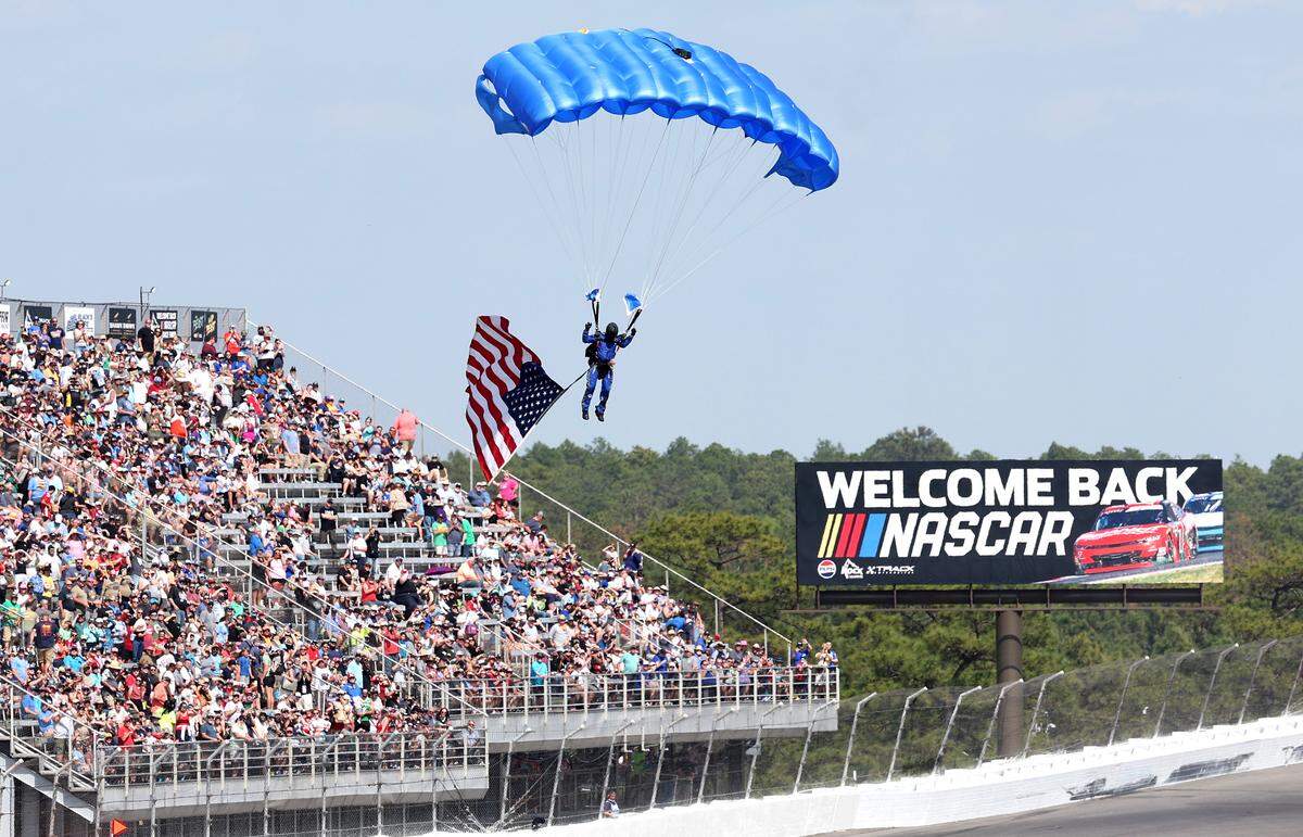 A paratrooper carries the American flag into the infield as NASCAR fans cheer at Rockingham Speedway on Saturday, April 19, 2025. The NASCAR Xfinity Series ran the North Carolina Education Lottery 250 at the speedway.