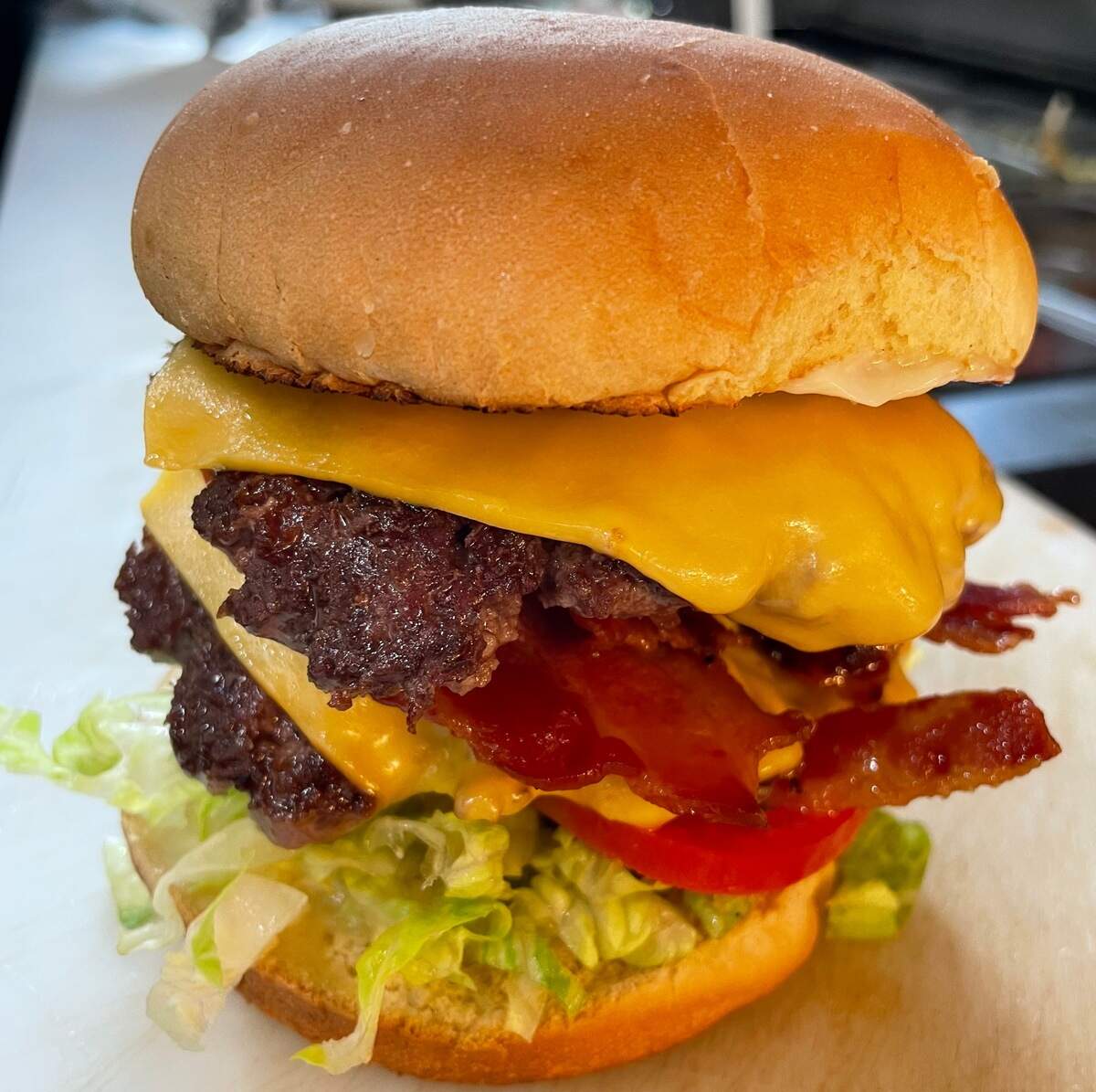 A detailed portrait shot of a double cheeseburger. From the bottom up, the burger has a bun, shredded lettuce, a slice of tomato, a seared beef patty with melted cheese, crispy bacon, a second seared patty with melted cheese, and a top bun.