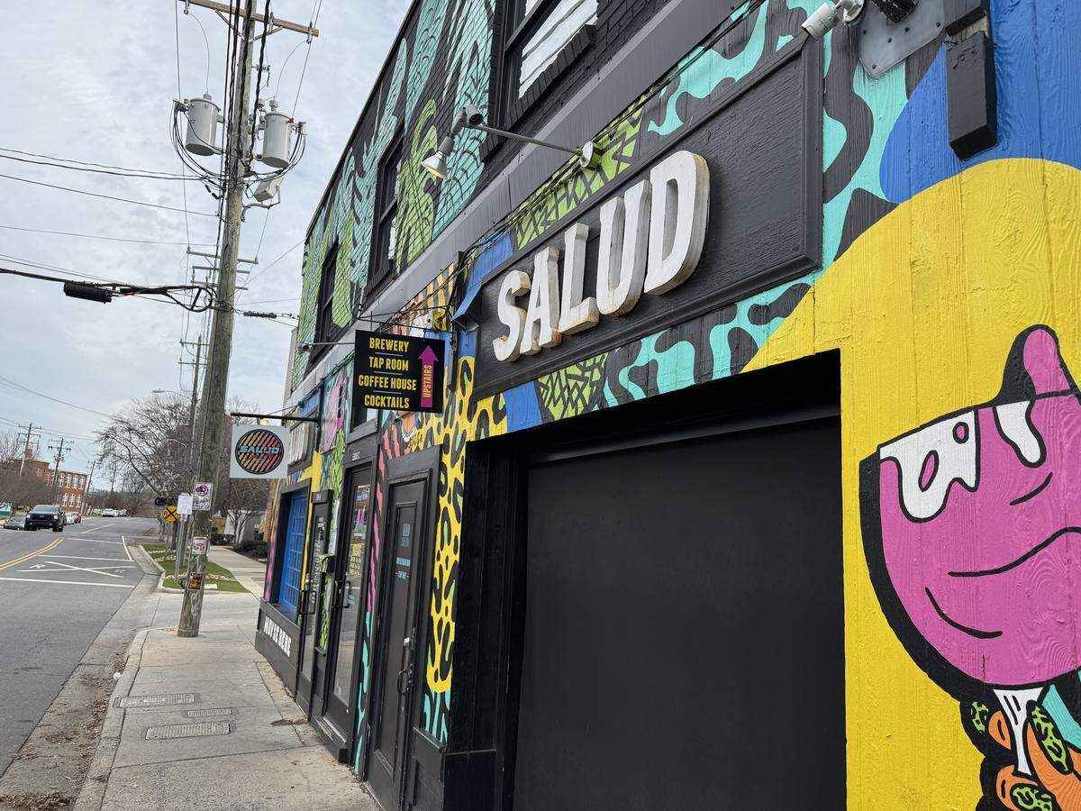A street-level view of a vibrant, two-story building featuring a massive, multi-colored pop-art mural. A large 3D sign reads “SALUD,” and smaller hanging signs identify the business as a brewery, taproom, coffee house, and cocktail bar.