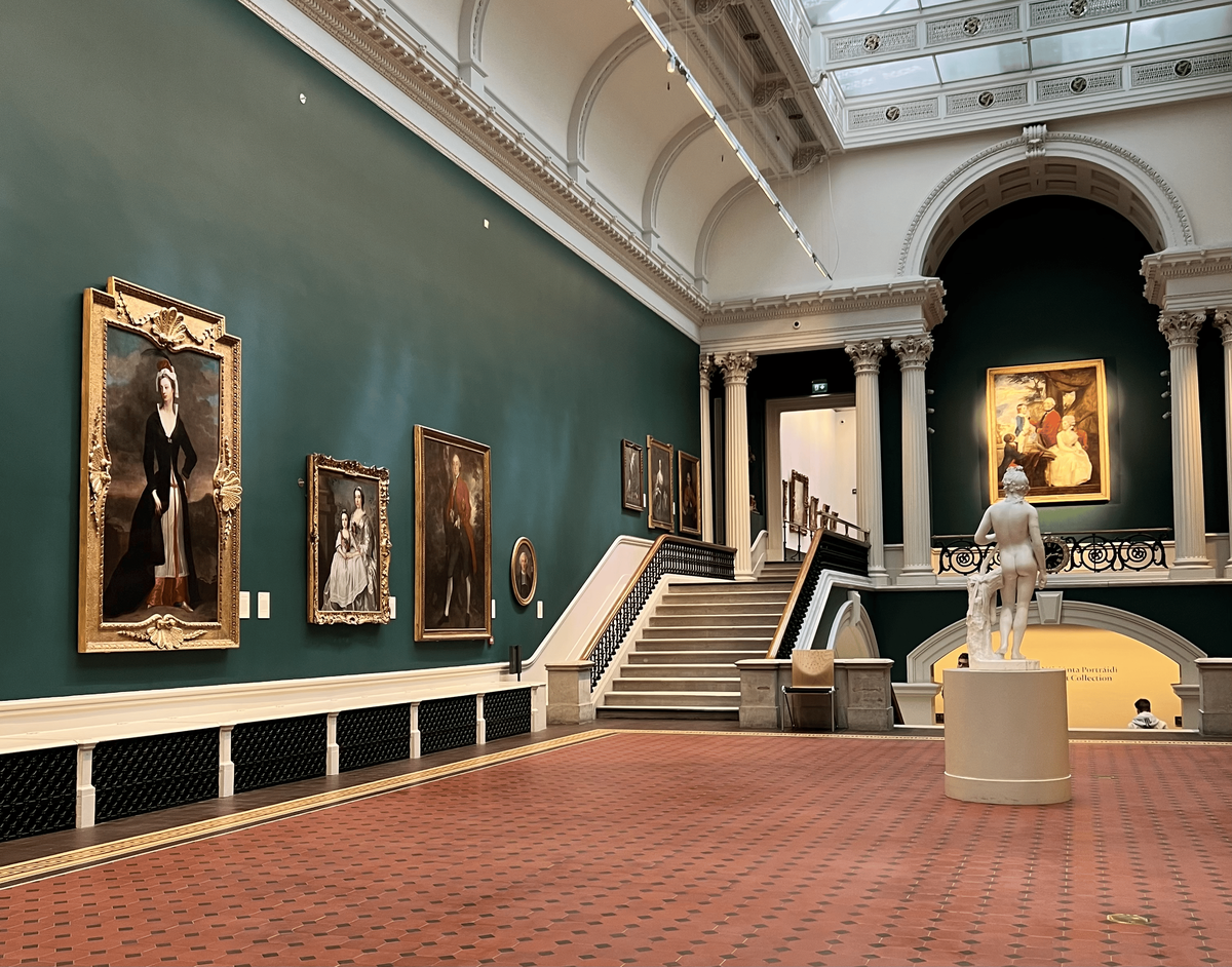 A large, ornate museum gallery with a high, vaulted ceiling and a terracotta-colored tiled floor. The walls are painted a deep green, and several large, gold-framed portraits are hung on them. In the center of the room, a grand staircase with a white railing leads to the upper level. On the right, a classical nude statue of a person on a white pedestal stands beneath a large arched alcove. Columns with Corinthian capitals support the upper level. The room is well-lit by natural light from a skylight and artificial lighting.